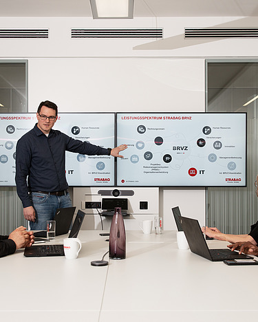 IT project management A man presents something to his team on two screens in a conference room.