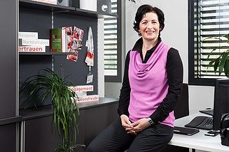 A woman in the office, sitting on the edge of a desk and smiling into the camera
