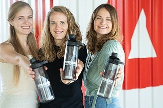 Three young women each hold a STRABAG water bottle up to the camera