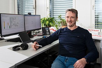 An IT employee sits at his desk and smiles at the camera.