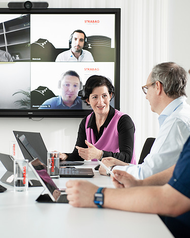 A woman presents something to her colleagues in a meeting. Four participants can be seen on a large screen in the background.