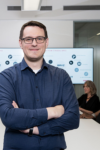 A young IT specialist with glasses looks into the camera. In the background, colleagues sit at a table and discuss things.