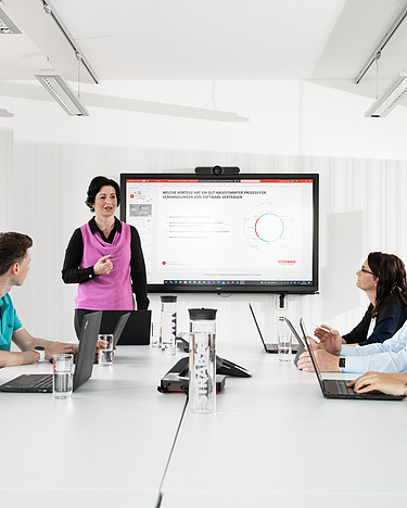 Eine Frau steht in einem hellen Konferenzraum vor dem Tisch mit ihrem Team.