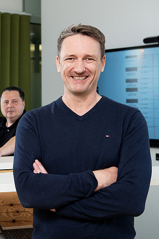 A standing man smiles at the camera. Three colleagues sit in the background and also look into the camera.