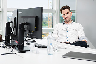 A man in a white shirt sits at a desk and looks into the camera.