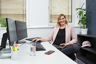 A young woman sits at an angle at a desk and smiles at the camera.