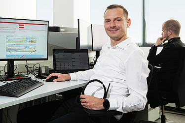 Ein ITler sitzt an einem Schreibtisch und blickt in die Kamera. Er hält ein Headset in der linken Hand.