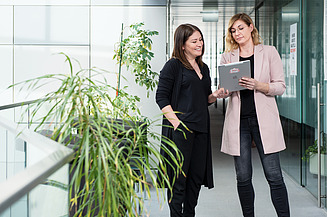 Your entry Two young women are standing in a bright office corridor looking at a tablet.
