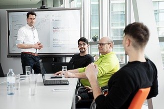 The IT project management group leader is in focus, while two colleagues are working on IT project manager tasks at the back. 
