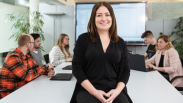 A colleague sits on a table and looks into the camera. In the background, a team sitting at a table is discussing something.