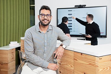 A young IT specialist with a beard and glasses looks into the camera. In the background, two colleagues are discussing a diagram on a screen.