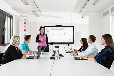 Eine Kollegin steht vor einem Bildschirm und blickt zu ihren Kolleg:innen an einem Konferenztisch.